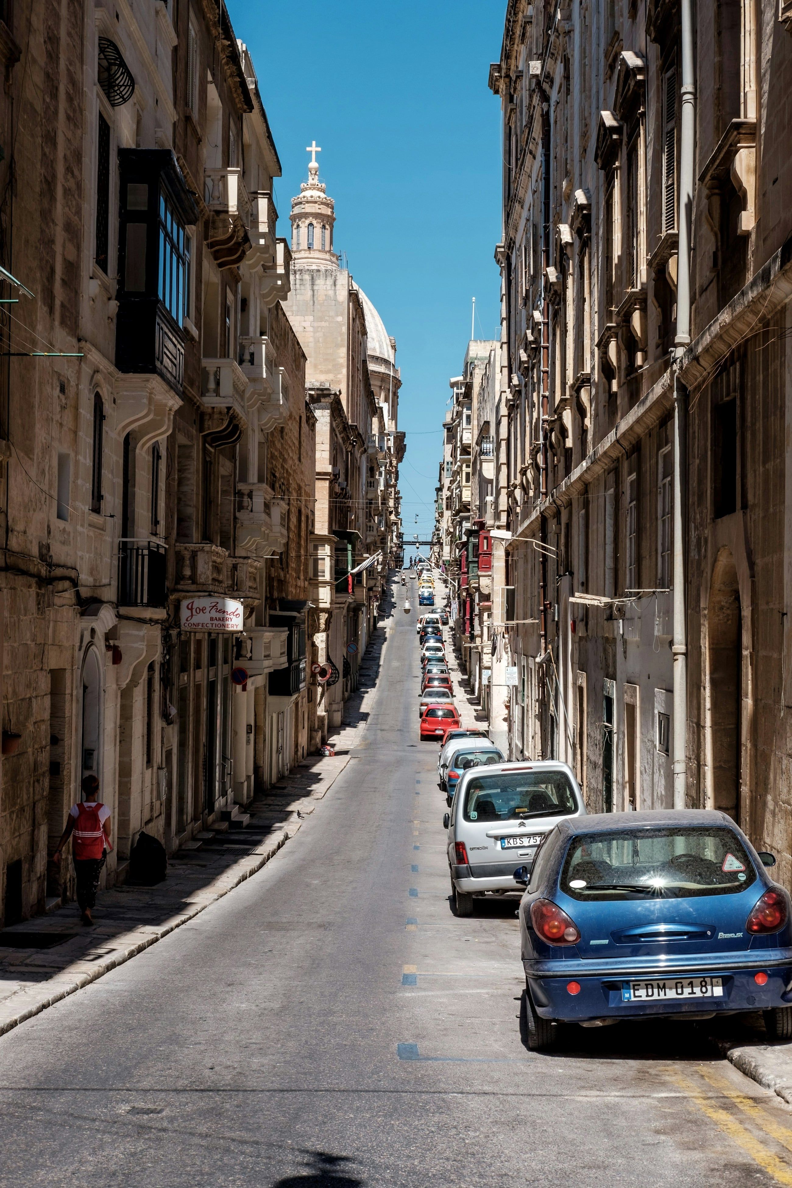 A steep Valletta street lined with parked cars and limestone balconies
